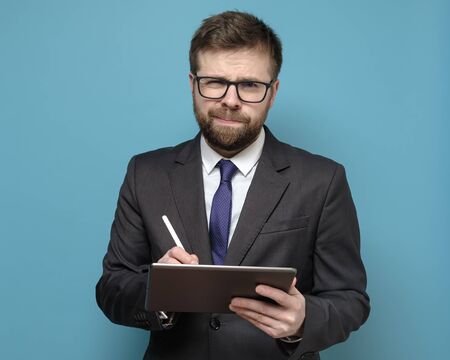 Bewildered businessman in a suit works with a tablet and stylus and looks thoughtfully at the camera with squinted eyes.の写真素材