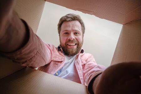Smiling Caucasian man unpacks a delivered box with a parcel or a gift, reaches out and takes the order. Unboxing inside view.の写真素材