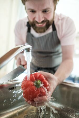 Male hand holds red pepper under a stream of water, and in another knife. Man in an apron is preparing for cooking, in the kitchen.の写真素材