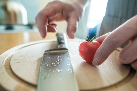 Female hands hold a red tomato on a cutting wooden board and are going to take a knife in droplets of water, in the kitchen.の写真素材