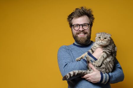 Funny smiling shaggy man hugs his adorable cat, who is not happy that the owner is holding it in hands. Copy space.の写真素材