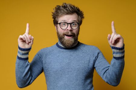 Great idea. Shaggy, bearded man with glasses and a blue sweater raises index fingers up and looks joyfully with mouth open.の写真素材