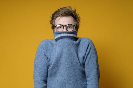 Shy, scared Caucasian shaggy man hiding behind his knitted sweater and looks uncertainly at the camera, on a yellow background.の写真素材