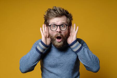 Surprised man with a shaggy hairstyle and beard listens to gossip and secrets, clutching hands to ears, on a yellow background.の写真素材