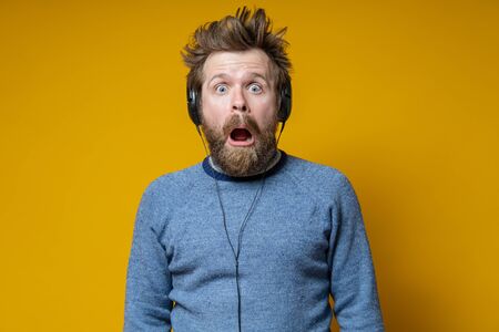 Man in the headphones is shocked. Shaggy music lover in an old sweater fearfully looks with his mouth open. Yellow background.の写真素材