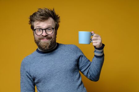 Attractive man with a shaggy hairstyle, glasses and an old sweater with a mug of hot drink has a good mood in the early morning.の写真素材