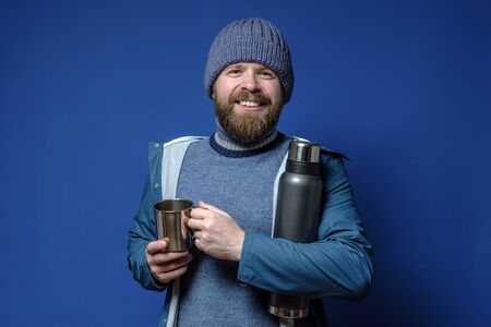 Joyful, smiling man with a thermos and a mug in hands, in a knitted hat and waterproof raincoat is preparing for the travel.の写真素材