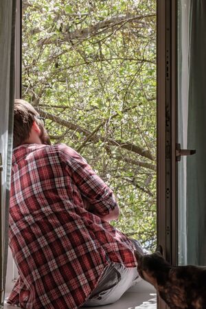 Man sitting on a windowsill at the open window and enjoying the cherry blossoms near his curious cat...