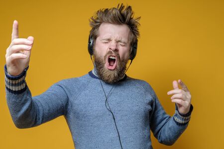 Satisfied hipster in an old sweater listens to music on headphones and loudly, emotionally sings along, making hand gestures.の写真素材