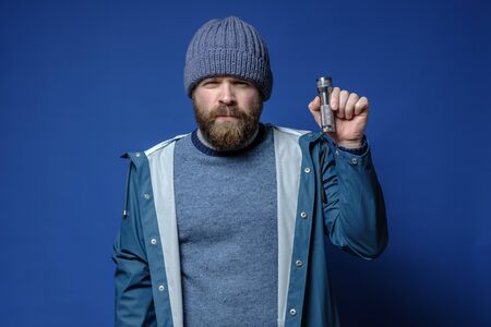 Serious bearded man in a knitted hat and a waterproof jacket holds a flashlight in hands, on a blue background.の写真素材