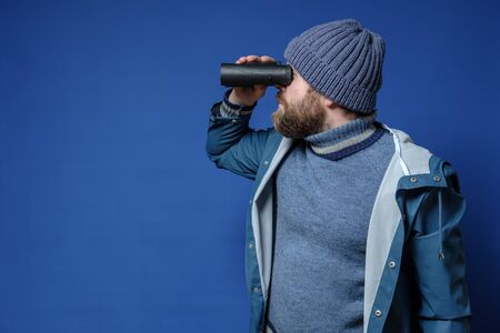 Bearded man in a knitted hat and a raincoat looks through binoculars, standing on an isolated blue background. Copy space.の写真素材