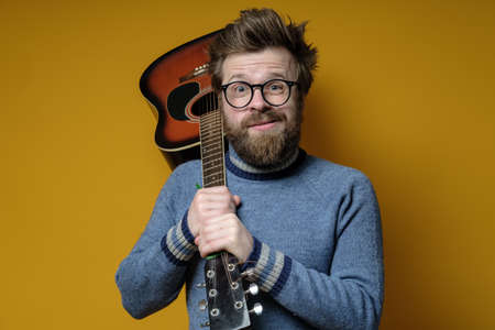 Funny hipster in an old sweater holds an acoustic guitar on shoulder and looks at the camera with a strange grimace.の写真素材