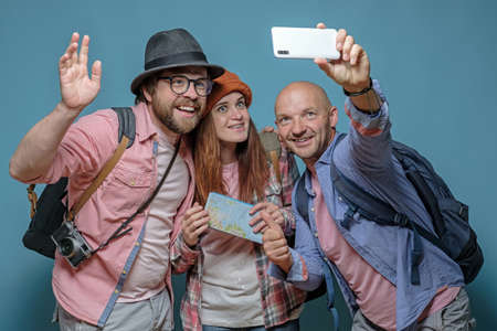 Three happy friends of tourists with backpacks travel and take pictures on a smartphone.