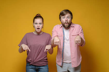 Surprised woman and man point down with their index fingers and stare wide-eyed at the camera, on a yellow background.の写真素材