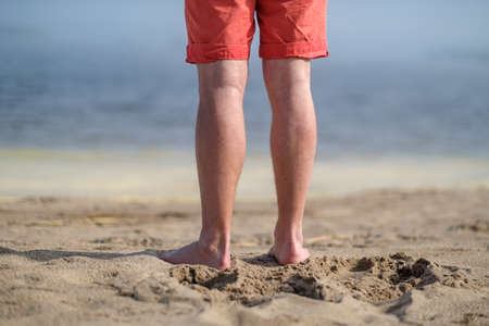 Barefoot male feet stand on a sandy beach, against a blurred ocean background, on a sunny day.の写真素材