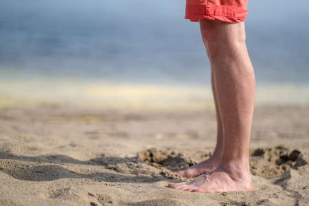 Barefoot male legs in shorts stand on a sandy beach, against a blurred ocean background, on a sunny, summer day. Copy space.の写真素材