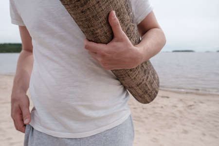 Man holds a yoga mat in hands, against the background of a sandy beach and a lake, on a summer day.の写真素材