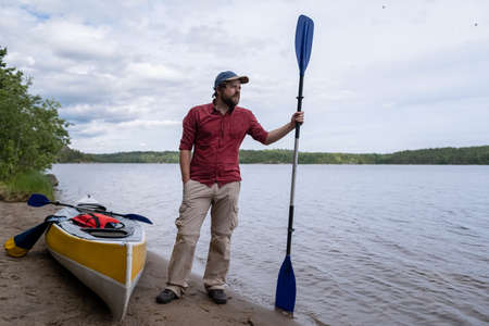 Attractive man with a paddle stands on a sandy beach, next to a kayak near the lake and looks into the distance. Lifestyle.の写真素材
