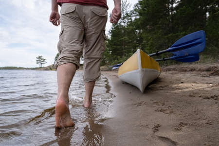 Barefoot male feet walk on the water, along the shore of the lake, against the background of a kayak with oars and trees. Lifestyle.の写真素材