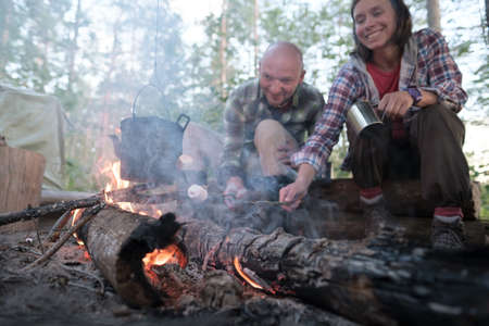 Happy friends fry marshmallows over a fire, over the flame of which a kettle hangs, a woman with a mug waiting for a hot drink.の写真素材
