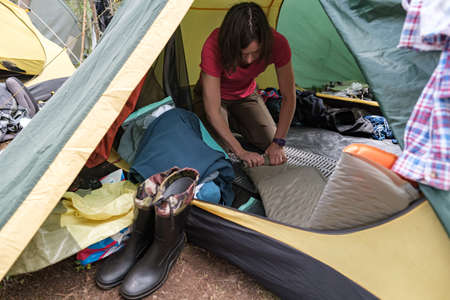 Focused tourist is twisting a camping inflatable mat while sitting in a tent. End of the hike, collection of things and equipment.の写真素材