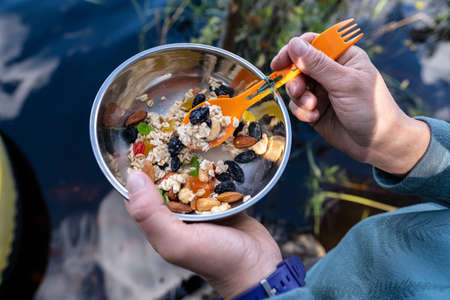 Hiking breakfast. Hands hold a metal plate in which delicious and healthy food, muesli, raisins,...