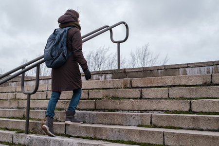 A tourist in warm clothes and a city backpack climbs the old stone steps of the stairs on a cloudy...