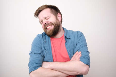 Funny shy man stands with crossed arms and smiles cute with eyes closed. White background.の写真素材