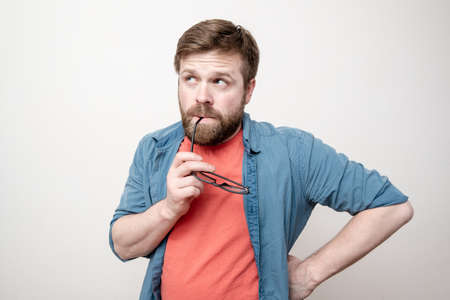 Pensive man holds glasses in mouth and tries to remember something, on a white background.の写真素材