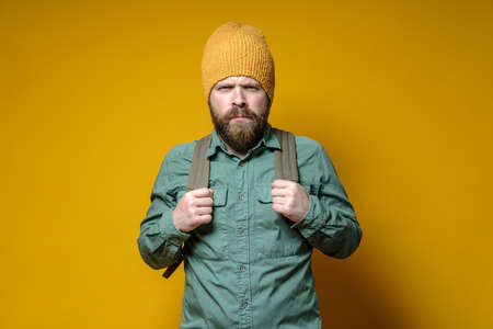 Angry bearded man in a knitted hat, shirt and with a city backpack frowns at the camera. Yellow background.の写真素材