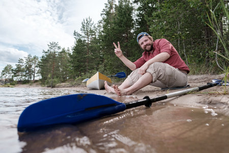 Joyful man is resting on the shore of the lake after kayaking, he will make a victorious gesture and smile while looking at the camera.の写真素材