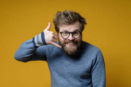 Funny shaggy-haired, bearded man wearing glasses and a sweater knitted making a hand gesture call me and looks camera. Yellow background.の写真素材