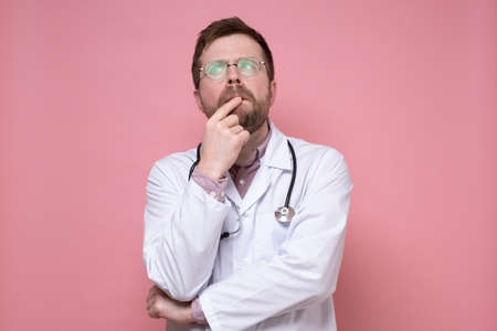 Puzzled male doctor with glasses, a white coat and a stethoscope around neck looks thoughtfully up. Pink background.の写真素材