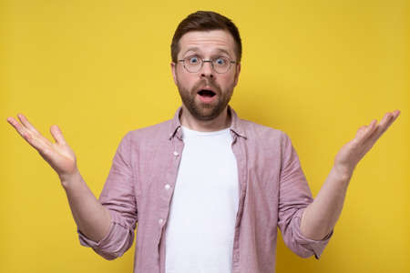 Shocked man in glasses and a shirt spreads arms to the sides and looks in bewilderment. Can not be. Yellow background.の写真素材