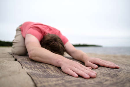 Man does yoga, he does the exercise in child pose, Balasana, relaxes and stretches lying on a rug, on a sandy beach by the lake.の写真素材