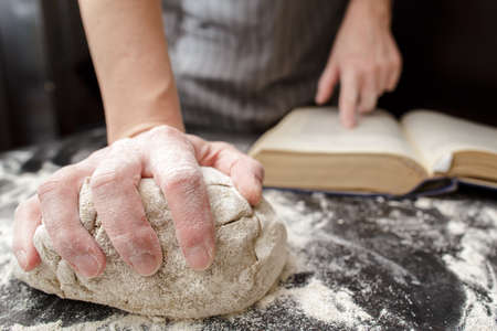 Baker holds one hand on the dough and the other on the cookbook, on the table sprinkled with flour. Bread making. Close-up.の写真素材