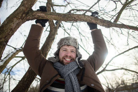 Funny, happy man holds hands to a tree branch, looks at the camera and smiles, against the background of the sky, on a spring day.の写真素材