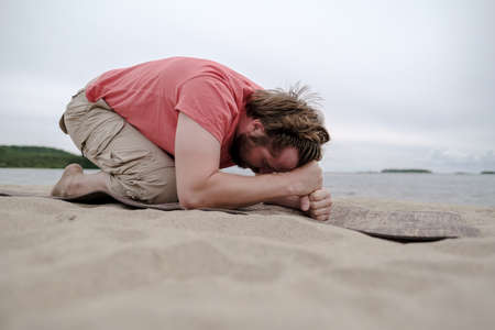 Calm man does yoga, he does the exercise, on the carpet, on the sandy shore of the lake. Healthy lifestyle concept.の写真素材