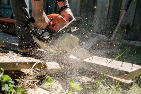 Male hands sawing old boards for firewood with a professional chainsaw, in a village yard. Winter preparation concept. Lifestyle.の写真素材
