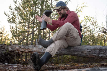 Man in hiking clothes sat on an old log in the woods, took off his rubber boots and shakes out trash. Active lifestyle concept.の写真素材
