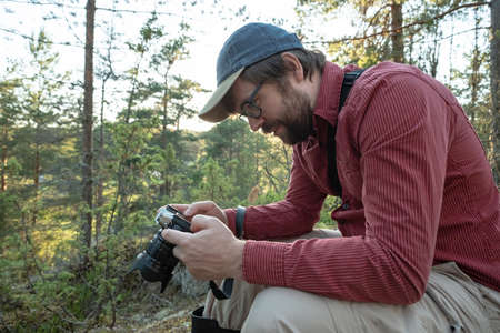 Photographer is resting, he holds a camera in hands and looks with interest at the photos taken while walking in the forest.の写真素材