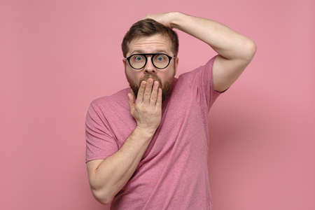 Shocked, strange man with glasses in fright covers mouth with one hand, put the other on head and looks with big eyes.の写真素材