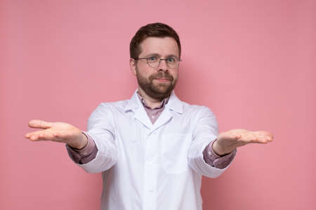 Friendly, charming doctor in a white coat makes a greeting gesture, open palms and looks into the camera. Welcome to the clinic.の写真素材