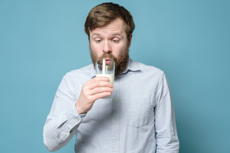 Lactose intolerance. A confused, bearded man pensively looks at a glass of milk in his hand. Blue background.の写真素材