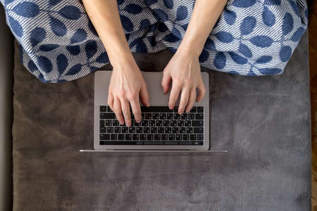 A woman wrapped in a plaid works with a laptop sitting on a soft sofa at home.の写真素材