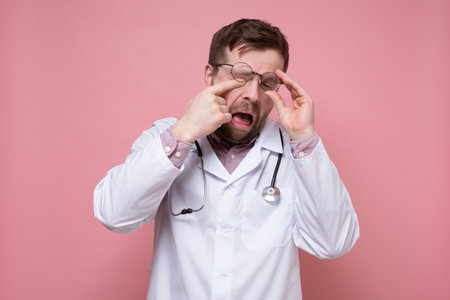 Tired, sleepy doctor with a stethoscope around neck raised glasses and rubs eyes. Pink background.の写真素材