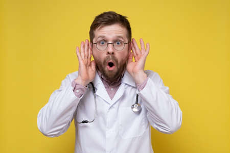 Shocked doctor has hands to ears and listens to secret conversations or gossip with an open mouth and big eyes.の写真素材