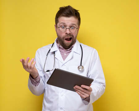 Young doctor with a stethoscope around neck holds a tablet in hands and looks at it in bewilderment. Yellow background.の写真素材