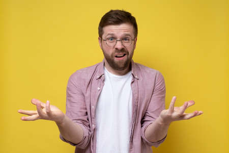 Perplexed Caucasian bearded man in glasses makes a gesture with hands and looks inquiringly into the camera. Yellow background.の写真素材
