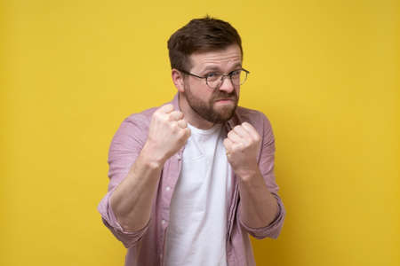 Aggressive bearded man with glasses clenched fists and started to defend himself with a funny expression on face. Yellow background.の写真素材